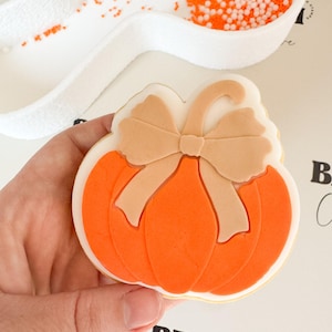 Puede incluir: Una galleta con forma de calabaza con glaseado naranja y un lazo marrón. La galleta se sostiene en una mano, con una cuchara blanca de chispas naranjas en el fondo. La galleta tiene un borde blanco.