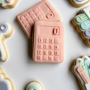 Könnte beinhalten: Zwei rosa Zuckerkekse, die wie Taschenrechner verziert sind. Die Kekse haben einen weißen Hintergrund und die Zahlen 0-9, Plus, Minus, Multiplizieren, Dividieren und Gleichheitszeichen sind in Schwarz gedruckt.