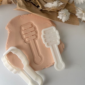 Könnte beinhalten: Ein weißer Ausstecher in Form eines Honiglöffels und ein Abdruck in rosa Knete. Ein zweiter weißer Honiglöffel-Ausstecher ist ebenfalls vorhanden. Trockenblumen und braunes Papier befinden sich im Hintergrund.