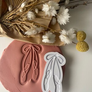 May include: White plastic cookie cutter shaped like a pair of ballet shoes with a bow on top. The cutter is on top of a pink clay surface.