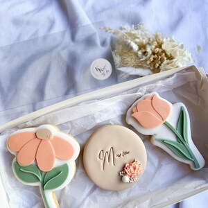 Puede incluir: Una caja de regalo llena de tres galletas decoradas. Las galletas tienen forma de flores y un c&iacute;rculo. La galleta circular tiene la palabra "Mum" escrita en ella. Las galletas est&aacute;n decoradas con glaseado rosa pastel y verde.