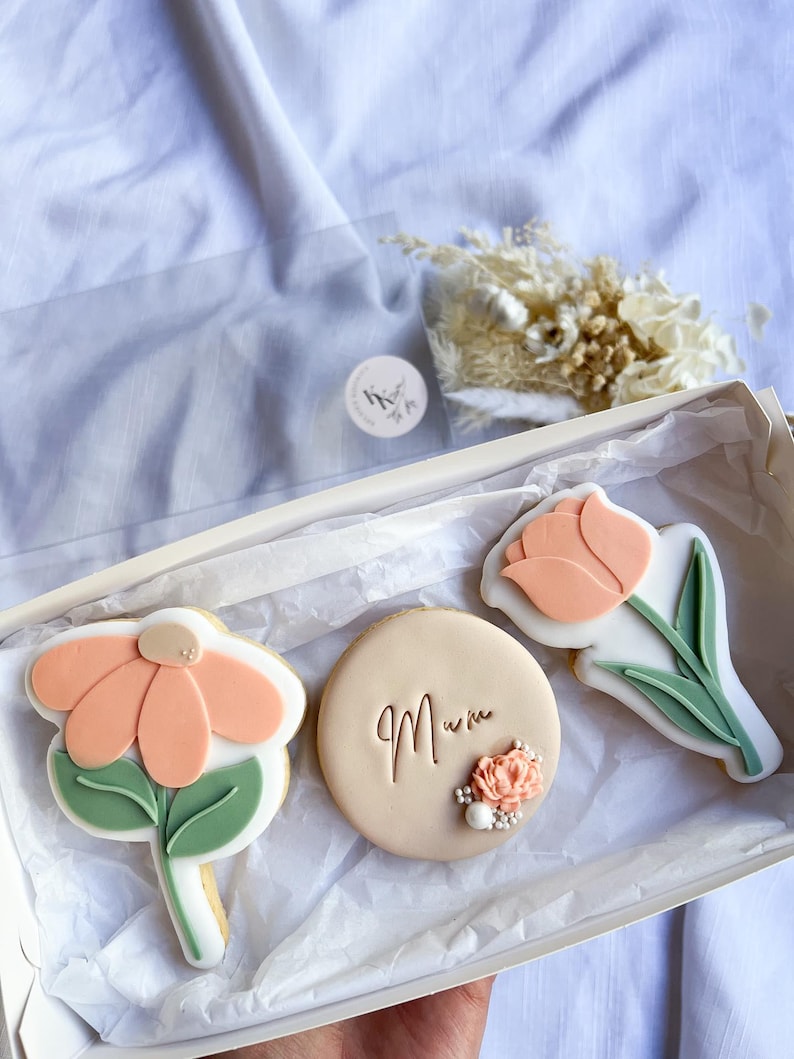 Puede incluir: Una caja de regalo llena de tres galletas decoradas. Las galletas tienen forma de flores y un c&iacute;rculo. La galleta circular tiene la palabra "Mum" escrita en ella. Las galletas est&aacute;n decoradas con glaseado rosa pastel y verde.