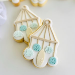 May include: Two decorated sugar cookies shaped like a mobile, with a white base and tan accents. The cookies are decorated with light blue seashell and sand dollar designs, and a surfboard. The cookies have a light yellow border.
