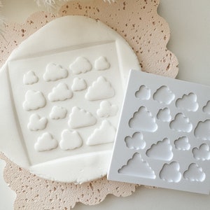 May include: A white silicone mold with multiple cloud shapes. The mold is shown next to a piece of white fondant with cloud shapes pressed into it.