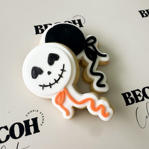 May include: Two Halloween-themed sugar cookies. One cookie is round with a white base, black facial features, and an orange bow. The other cookie is black and white with a bow. The cookies are on a white surface.