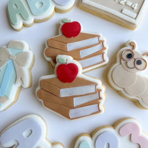 May include: A selection of decorated sugar biscuits with a school theme. Biscuits include the letters "ABC", a chalkboard with numbers, a stack of books topped with a red apple, a bow, and a caterpillar wearing glasses.