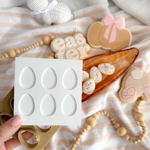 Puede incluir: Un surtido de artículos con temática de Pascua, que incluyen una figura de conejito blanco, un molde con forma de huevo y galletas decoradas. Las galletas presentan un lazo, patas de conejo y huevos moteados. También hay una guirnalda de cuentas de madera y una bandeja con huevos moteados.