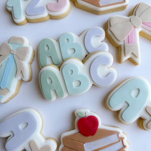 Könnte beinhalten: Ein Set aus neun dekorierten Zuckerkeksen mit Schulthema. Die Kekse sind in Pastellfarben verziert und beinhalten Zahlen, Buchstaben, einen Bücherstapel, einen Apfel und eine Schleife.