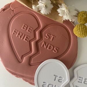 May include: Two pink heart-shaped cookie cutters with the words "BE FRIENDS" and "BEST FRIENDS" imprinted on them. The cutters are broken in half, with the words split between the two pieces.