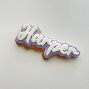 May include: A purple and white cookie decorated with the name "Harper" in white icing.