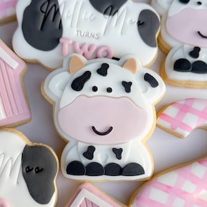 Puede incluir: Surtido de galletas de azúcar decoradas con temática de granja. Las galletas incluyen una vaca, un granero y una galleta con el texto "Millie Moo Turns Two". Las galletas son blancas, rosas, negras y beige.