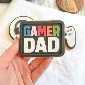 May include: A decorated cookie with the words "GAMER DAD" in colorful letters on a black background. The cookie is held in a hand, and there are other gaming-themed cookies and a headset in the background.