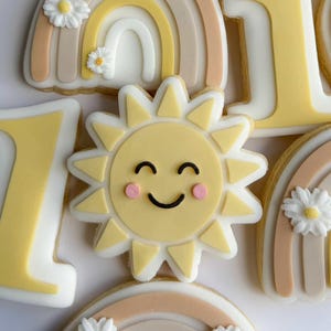 Peut inclure: Assortiment de biscuits décorés sur le thème du soleil. Les biscuits présentent un soleil souriant, des arcs-en-ciel, des marguerites et le chiffre 1, dans des tons pastel. Ces biscuits sont probablement pour une fête d'anniversaire.
