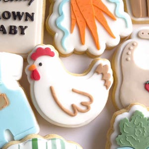 Puede incluir: Surtido de galletas de az&uacute;car decoradas. Una galleta de pollo blanco con cresta y pico rojos est&aacute; rodeada de otras galletas, incluyendo un babero con fresas, zanahorias y un traje de beb&eacute;. Las galletas est&aacute;n decoradas con glaseado blanco, naranja, azul y marr&oacute;n.
