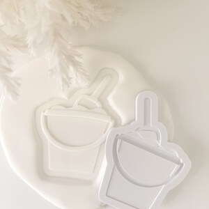 May include: Two white plastic cookie cutters shaped like buckets with handles. The cutters are on a white surface.