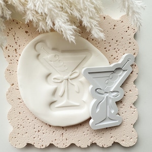 May include: White silicone mould of a martini glass with olives and a bow. The mould is on a textured surface.