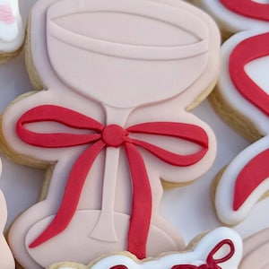 May include: Close-up of decorated sugar biscuits. The biscuits are shaped like a wine glass with a red bow. Other biscuits are visible, including a white biscuit with a red ribbon design. The biscuits are on a white surface.