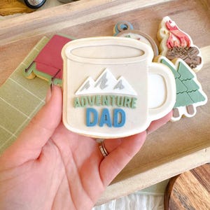 May include: A beige cookie shaped like a mug with the words "ADVENTURE DAD" in blue and green. The mug design includes mountain graphics. Other cookies in the image include a tent, a campfire, and a Christmas tree.