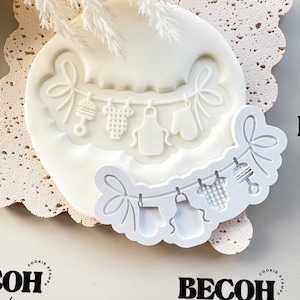 May include: A white silicone cookie stamp with a baby shower design. It features a banner with baby clothes, a rattle, and bows. The stamp has the text "BECOH Collective" in black.