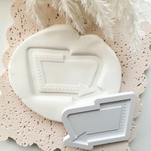 Può includere: Due stampi in silicone bianco per la cottura o l'artigianato. Gli stampi sono a forma di cartelli retrò con una freccia che punta a destra. Gli stampi hanno un bordo punteggiato e una freccia in rilievo.