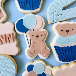 Può includere: Assortimento di biscotti di zucchero decorati nei toni del blu, rosa e bianco. I disegni includono un orsetto con palloncini, cupcake e il numero "2". I biscotti sono disposti su una superficie azzurra.
