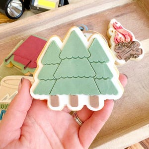 Könnte beinhalten: Eine Hand hält einen grünen, baumförmigen Keks mit hellbraunem Boden und weißem Zuckerguss-Umriss. Weitere Kekse zum Thema Camping und eine Taschenlampe befinden sich im Hintergrund. Die Kekse liegen auf einem Holztablett.