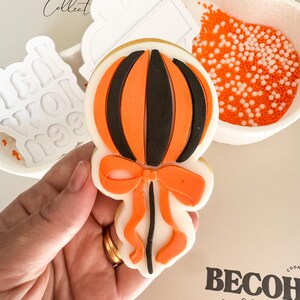 May include: A decorated cookie in the shape of a pumpkin with orange and black stripes and a bow. The cookie is held in a hand, with other cookie cutters and orange sprinkles in the background. The text "BECOH Collective" is visible.