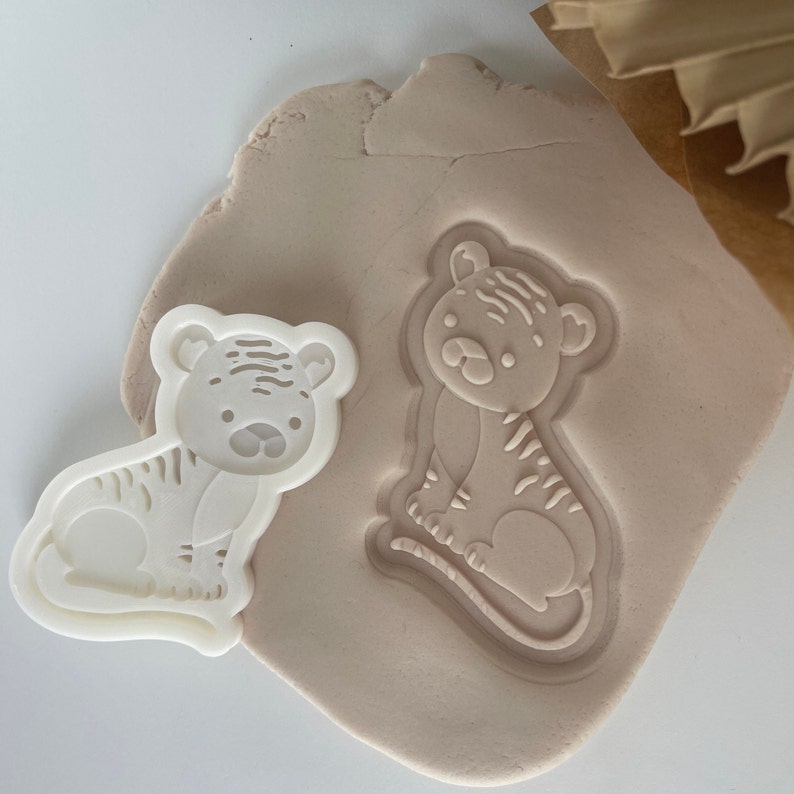 May include: White plastic cookie cutter in the shape of a tiger, with a tiger imprint in brown clay.
