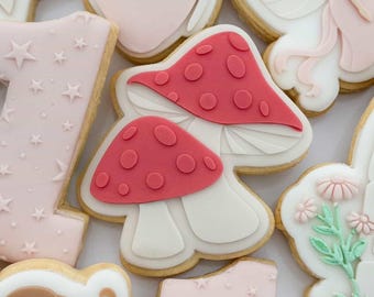 Op de afbeelding: Assortiment van versierde suikerkoekjes met een lichtroze en wit kleurenschema. De koekjes hebben ontwerpen zoals paddenstoelen, sterren, een fee, een vlinder en het nummer één. De koekjes hebben een lichtbruine rand.