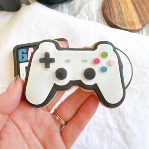May include: A white cookie in the shape of a video game controller, outlined in black, with colourful buttons. The controller features a black cross and two black circles. A second cookie with the word "GAME" is partially visible. Headphones and a wooden coaster are in the background.