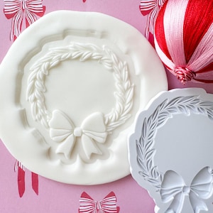 May include: Two white silicone molds for baking. One mold is a wreath with a bow in the center. The other mold is a wreath with a bow at the bottom.