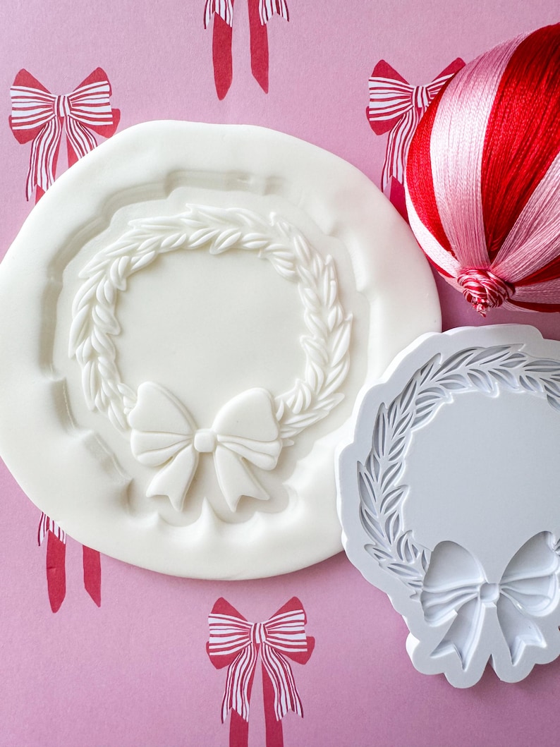 May include: Two white silicone molds for baking. One mold is a wreath with a bow in the center. The other mold is a wreath with a bow at the bottom.