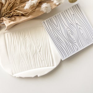 May include: A white, square silicone mold with a wood grain pattern. A piece of white clay is pressed with the mold, showing the detailed wood texture. Dried flowers and foliage are in the background.