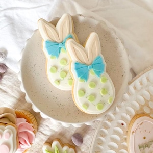 May include: Two Easter bunny-shaped biscuits with light blue bows and green polka dots on a white plate. Other Easter-themed biscuits, including a carrot and a flower, are also visible.
