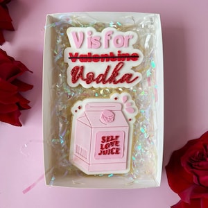 May include: Two pink and red decorated cookies in a white box. The top cookie says "V is for Vodka" with the word "Valentine" crossed out. The bottom cookie is shaped like a milk carton and says "Self Love Juice".