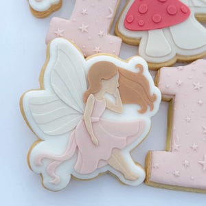 以下が含まれることがあります： デコレーションされたクッキーのクローズアップ。白い翼とピンクのドレスを着た妖精のクッキーは、数字の1とキノコを含む他のクッキーに囲まれています。クッキーはピンク、白、茶色のアイシングで飾られています。
