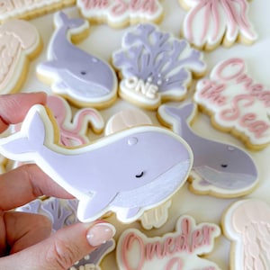 Puede incluir: Una colección de galletas de azúcar decoradas con temática marina. Las galletas presentan diseños de ballenas, corales, medusas y pulpos en colores pastel. Algunas galletas tienen el texto "One" y "Under the Sea".