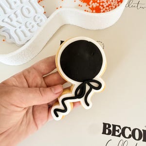 May include: A decorated sugar cookie in the shape of a balloon, with a black center and white border. The balloon has a black bow and string. The cookie is held in a hand, with a white tray of orange and white sprinkles in the background.