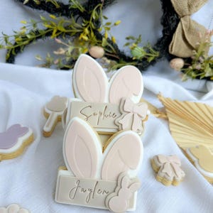 Peut inclure: Deux biscuits en sucre décorés avec des oreilles de lapin et des noms personnalisés. Le biscuit du haut dit "Sophie" et le biscuit du bas dit "Jaylen".