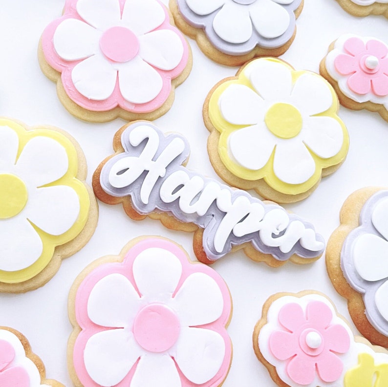 May include: A set of decorated sugar cookies with a daisy design. The cookies are decorated with white, yellow, pink, and purple icing. One cookie has the name "Harper" written in purple icing.