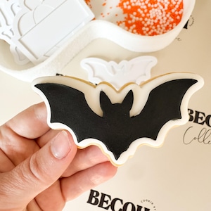 May include: A black bat-shaped cookie with white and gold accents is held in a hand. White cookie cutters and orange sprinkles are in the background. The text "BECOH Collective" is visible.
