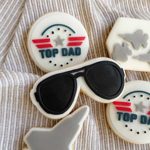 Puede incluir: Galletas de azúcar decoradas con el tema "Top Dad". Las galletas presentan diseños de gafas de sol de aviador, aviones de combate y las palabras "Top Dad" en azul, rojo y blanco. Perfectas para el Día del Padre o una fiesta temática.