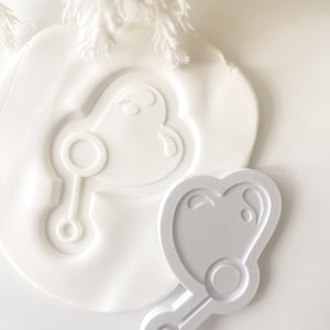 May include: White plastic cookie cutters shaped like a rattle, one with a heart-shaped handle and the other with a round handle.