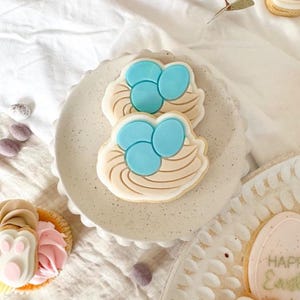 May include: Two decorated sugar cookies shaped like bird nests with blue eggs. The cookies are on a white plate with a speckled pattern.