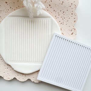 May include: A white, rectangular silicone mold with vertical lines, next to a white, textured clay piece with a similar pattern. The clay piece is on a textured, light pink surface. White decorative sprigs are in the background.