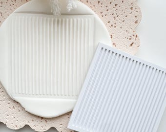 Puede incluir: Un molde de silicona blanco rectangular con líneas verticales, junto a una pieza de arcilla blanca texturizada con un patrón similar. La pieza de arcilla está sobre una superficie rosa claro texturizada. Ramitas decorativas blancas en el fondo.