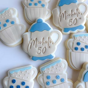 Puede incluir: Galletas de azúcar decoradas con forma de tazas de té y teteras. Las galletas son blancas con detalles azules y diseños florales. Algunas galletas tienen el texto "Michele's 50th".