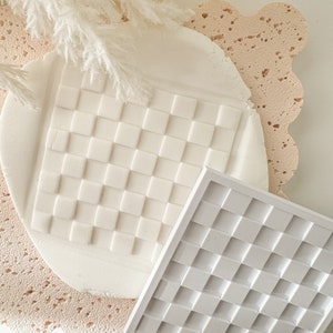 Può includere: Due strumenti per la decorazione di torte in silicone bianco con un motivo a griglia quadrata. Gli strumenti sono su una superficie rosa testurizzata.
