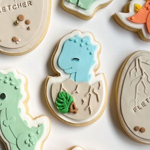 May include: A set of five decorated sugar cookies featuring a dinosaur theme. The cookies are decorated with green, blue, and brown icing. One cookie features a blue dinosaur hatching from an egg.