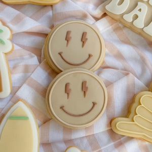 Puede incluir: Surtido de galletas de azúcar decoradas con temática playera. Dos galletas redondas presentan una carita sonriente con ojos de rayo. Otras galletas incluyen una tabla de surf, un gesto con la mano y una palmera. Las galletas están sobre un paño a rayas color melocotón.